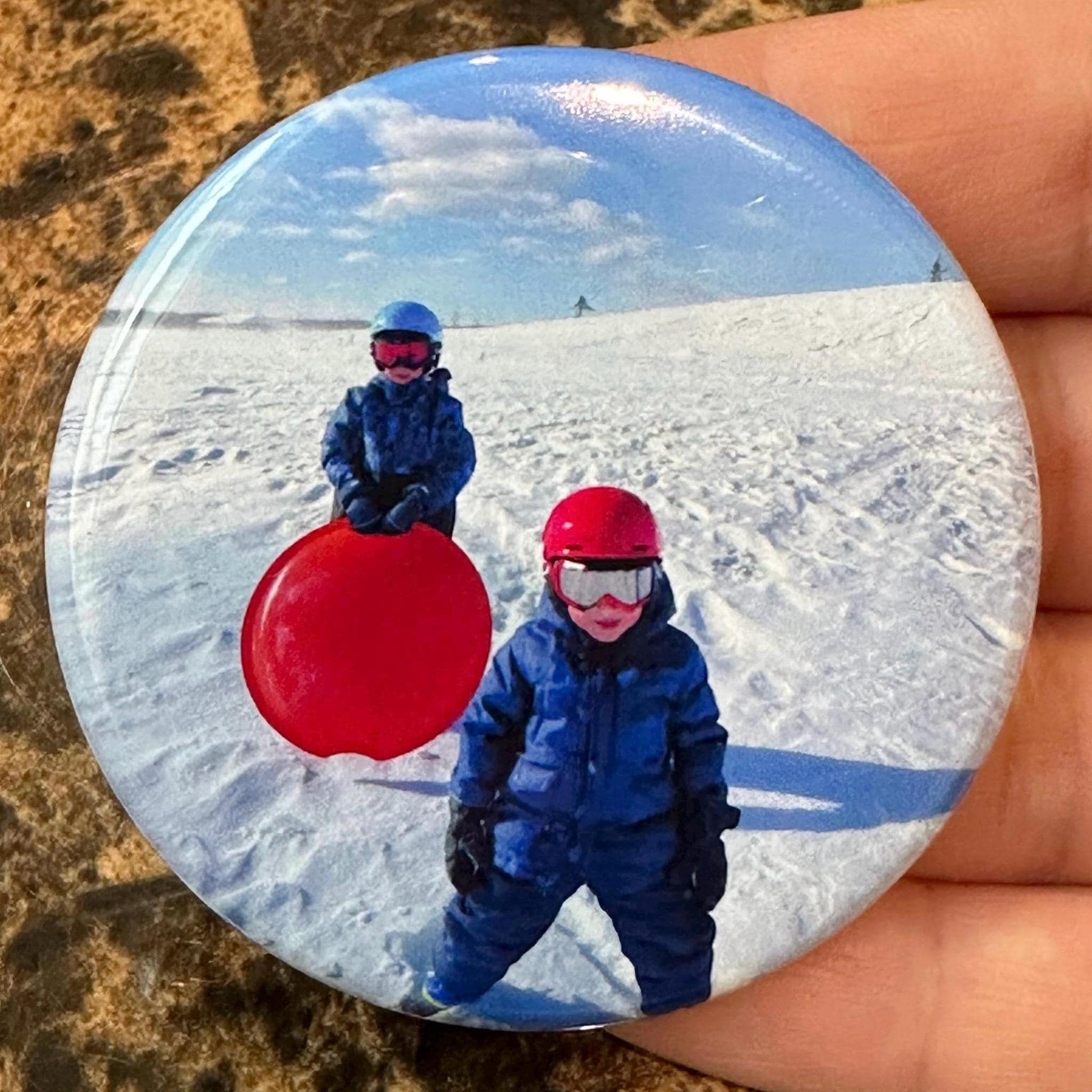 Button magnet with two children in winter clothing and a red sled on a snowy background