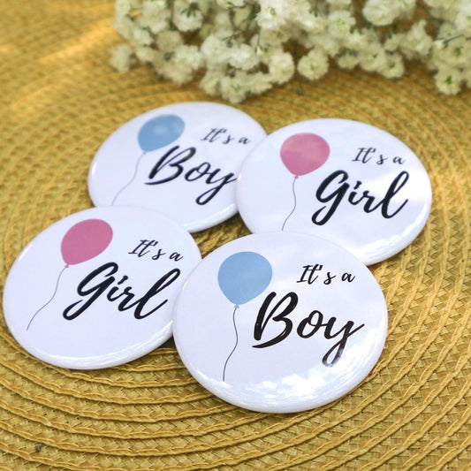 Four buttons with 'It's a Boy' and 'It's a Girl' text and balloon graphics on a textured surface. Gender reveal pins
